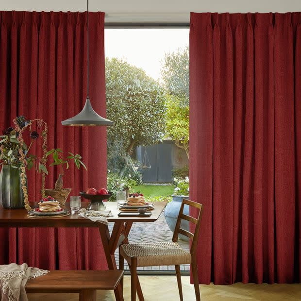 Deep red pleated floor-length curtains, split centrally and textured; partially drawn, framing and filtering daylight; dining room with a table set under a pendant lamp, opening to a green garden.