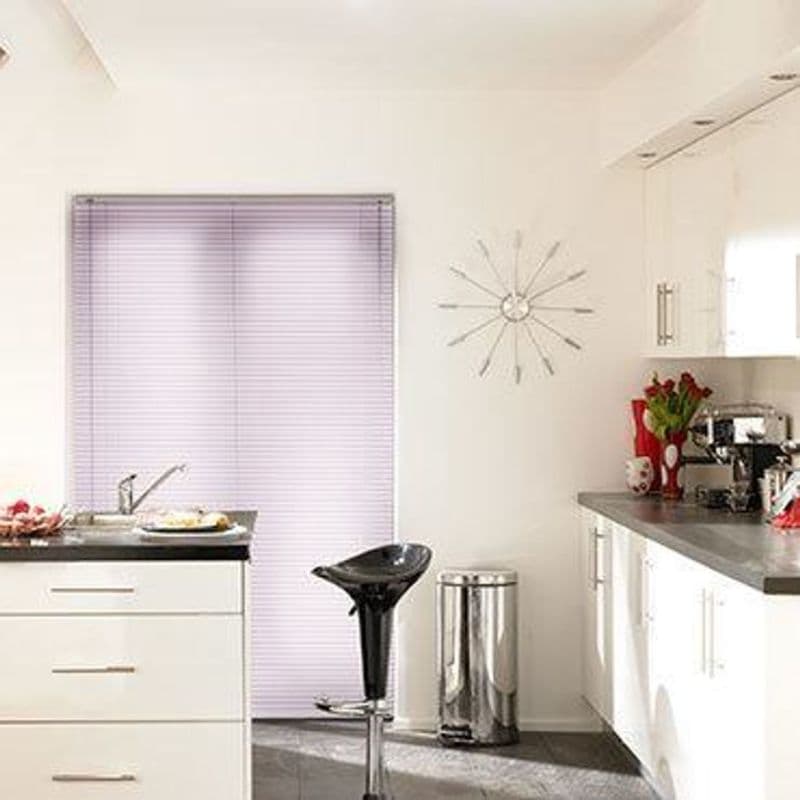 Horizontal venetian blinds covering a sliding door, fully closed and softly filtering daylight; modern white kitchen setting with black countertop and stool, stainless bin, sunburst wall clock and red tulips.