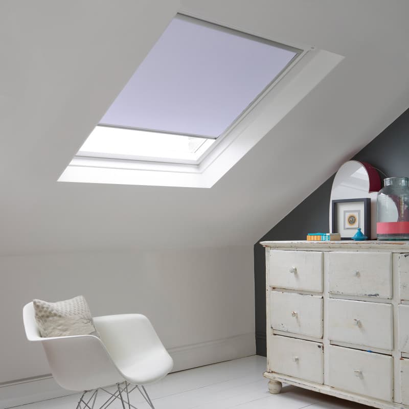 Room with white eaves wall, dark blue-grey wall to right with a shabby chic white drawer unit, a contemporary white chair, white painted floorboards and a skylight blind in pale lilac.