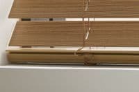 Wooden horizontal slatted blinds, lowered and slightly tilted with cords looped and hanging; blocking most light through a white window frame, interior wall and windowsill visible.