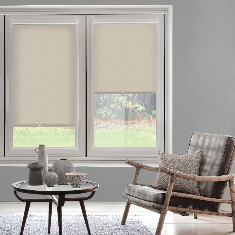 Beige roller blinds over twin windows, partially lowered with clear lower panes, filtering soft daylight into a gray-walled living room with a wooden armchair, patterned cushion and round coffee table.