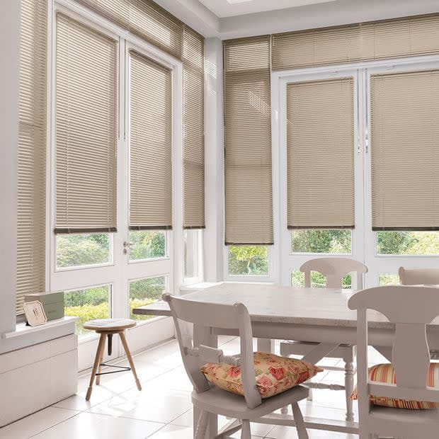 Beige horizontal Venetian blinds covering tall white-framed windows and French doors, lowered and filtering daylight; sunlit dining area with white wooden table, chairs with floral cushions, tiled floor, garden view.