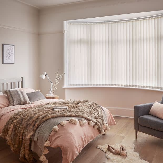 Cream vertical blinds cover a bay window, closed and gently diffusing daylight. In a soft-neutral bedroom a double bed with textured throws, bedside lamp, wooden floor and a gray armchair.