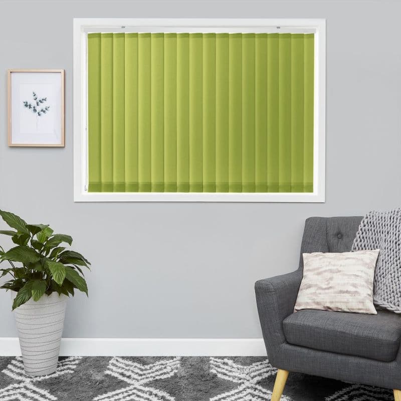 Green vertical blinds, fully closed across a white-framed window with evenly aligned slats; blocking light and creating uniform color. Context: grey-walled living room with potted plant, framed print, grey armchair.
