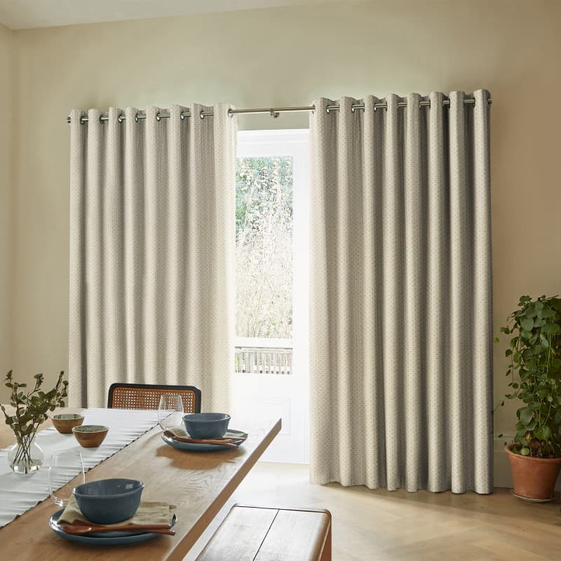 Grommet-top beige patterned curtains spanning a doorway on a metal rod, partially drawn filtering bright daylight; dining room with a wooden table set with bowls and a potted plant by the wall.