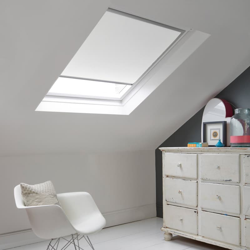Room with white eaves wall, dark blue-grey wall to right with a shabby chic white drawer unit, a contemporary white chair, white painted floorboards and a skylight blind in white.