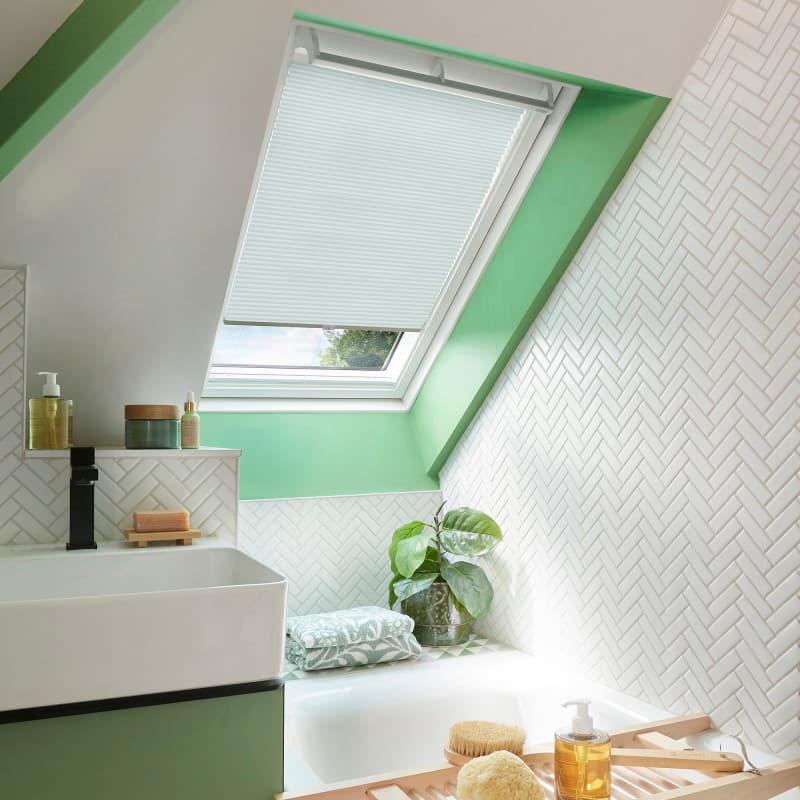 White pleated skylight blind, lowered and filtering soft daylight; set in a green-trimmed attic bathroom with white herringbone tiles, a sink and tub edged with folded towels, plant, and toiletries.