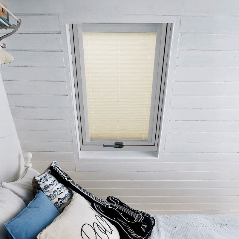 Beige pleated blind mounted within a white-framed skylight, fully lowered and diffusing soft daylight; attic bedroom with slanted white wood paneling and a bed topped with blue and patterned pillows beneath.