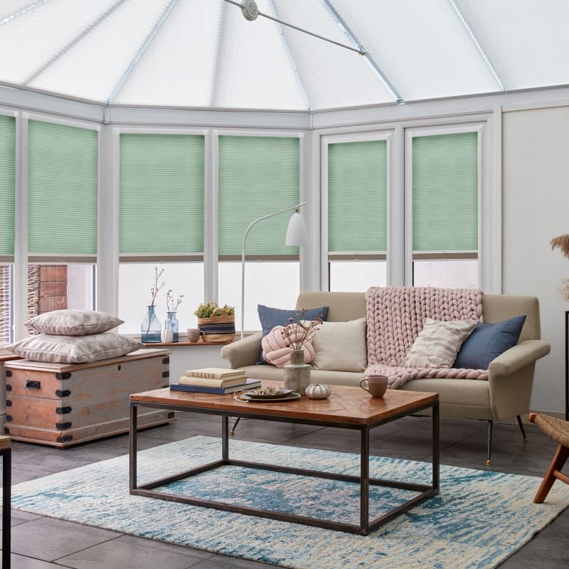 Mint-green cellular blinds lowered halfway across white-framed conservatory windows, filtering soft daylight; cozy sunroom living area with beige sofa, chunky pink throw, wooden coffee table and blue rug.