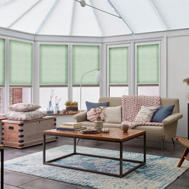 Light-green pleated cellular blinds, lowered halfway across tall white-framed conservatory windows, gently filtering soft daylight; a cozy living area with beige sofa, wooden coffee table, textured rug and plants.