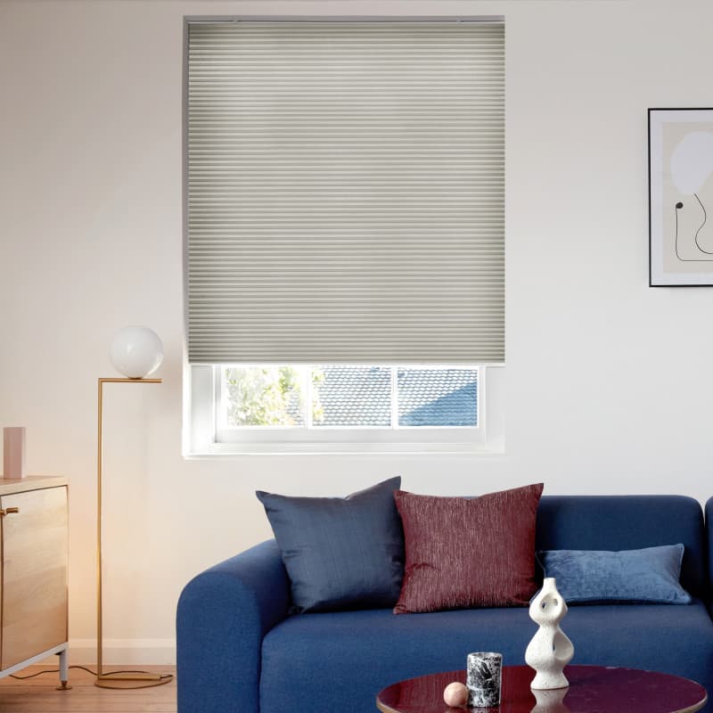 A light-gray pleated cellular blind, lowered with subtle horizontal ribs and a bottom gap, filtering daylight in a bright living room with blue sofa, cushions, floor lamp and coffee table.