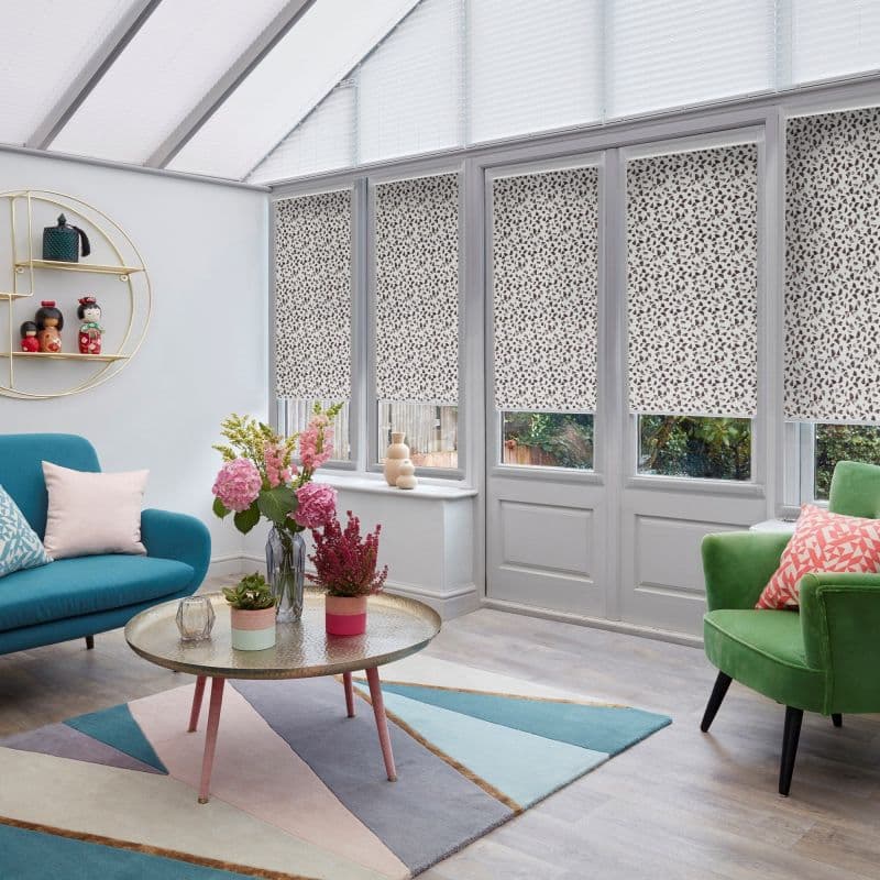 Patterned roller blinds (white with small dark leaf-like speckles) lowered over tall conservatory windows and door, softly filtering daylight; bright living area with teal sofa, green armchair and floral centerpiece.