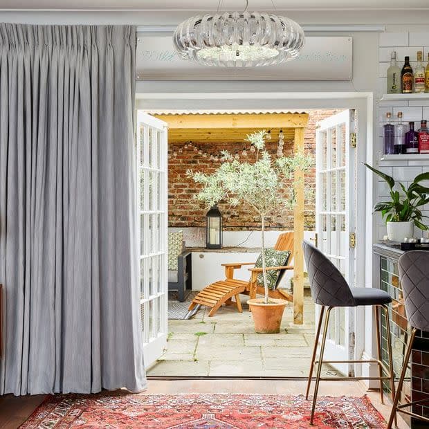 Grey floor-length pleated curtains pulled aside, revealing open white French doors to a living area with bar stools, opening onto a patio with wooden lounge chair, potted tree, brick wall.