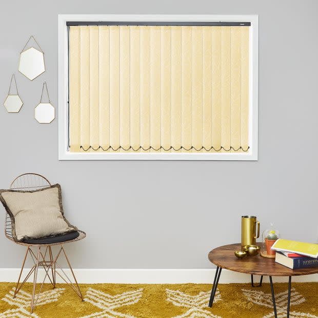 Beige textured vertical blinds, fully closed with scalloped weighted hems and bottom chain, filtering soft daylight through a white-framed window; gray-walled living room with wicker chair, coffee table, mustard rug.