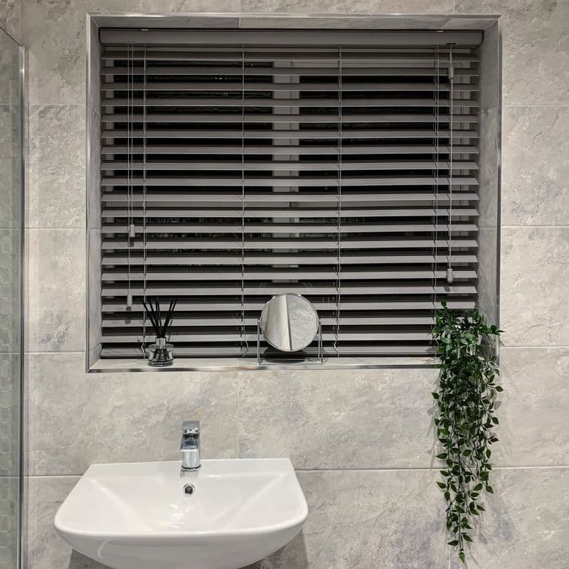 Grey horizontal venetian blinds, fully closed with visible cords and slats, filtering dim light through a recessed tiled bathroom window; white sink, small round mirror, diffuser, trailing plant on sill.