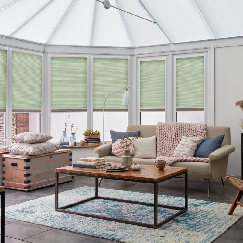 Pale green cellular blinds cover conservatory bay windows, lowered and filtering daylight; a neutral sofa with chunky pink throw and wooden coffee table sit on a blue rug.