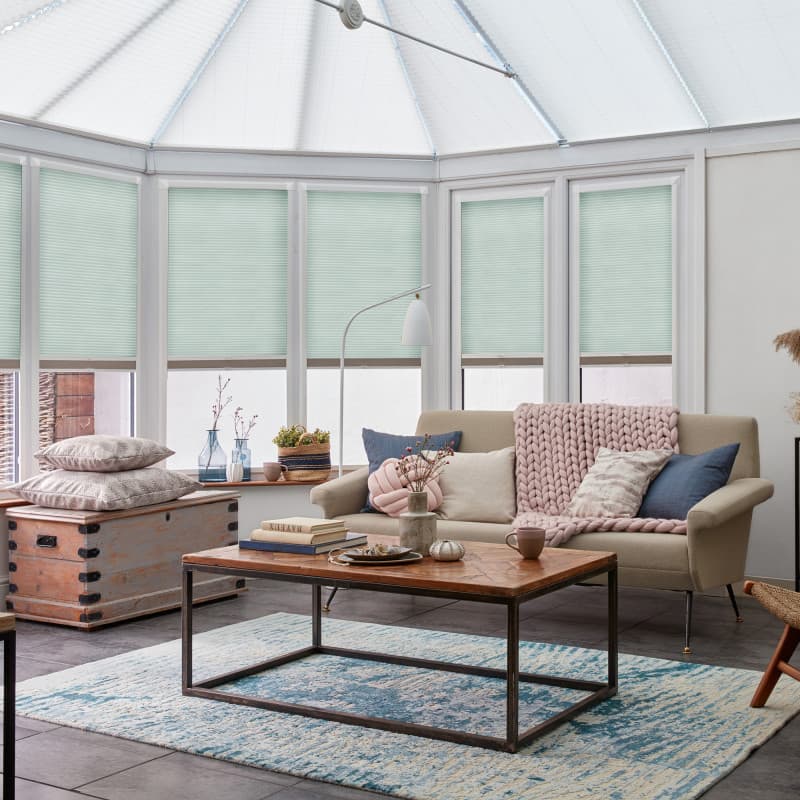 Mint-green cellular (pleated) blinds covering tall conservatory windows, lowered to just above the sills and filtering soft daylight into a cozy sunroom with a sofa, chunky knit throw, and wooden table.
