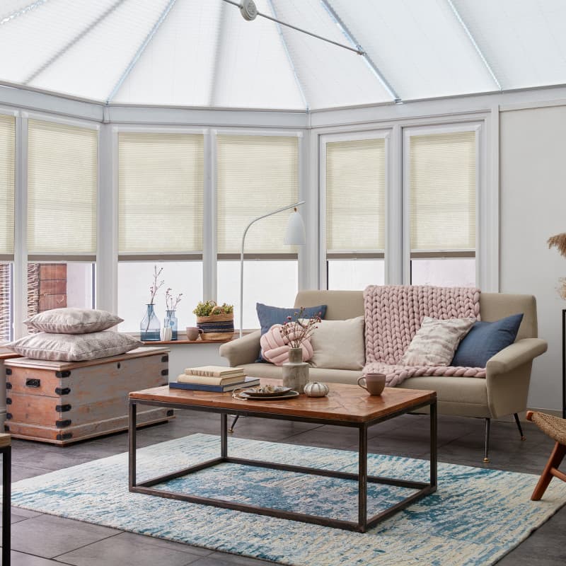 Beige pleated cellular blinds lowered mid-window, filtering daylight; across a bay of tall windows beneath a glazed roof, framing a bright conservatory living room with sofa, coffee table and blue rug.