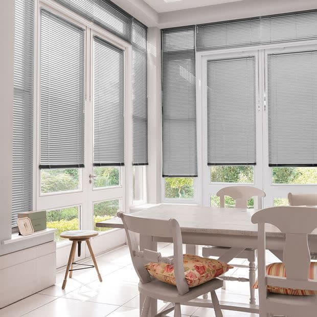Horizontal aluminium Venetian blinds on tall French windows and doors, lowered with slats slightly tilted to filter daylight; sunlit dining nook with wooden table and chairs, patterned cushions, garden view.