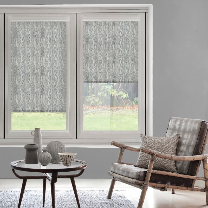 Roller blinds with a subtle vertical wave pattern, left fully lowered and right halfway, filtering daylight; in a grey-walled living room with a rattan armchair, coffee table and lawn view.