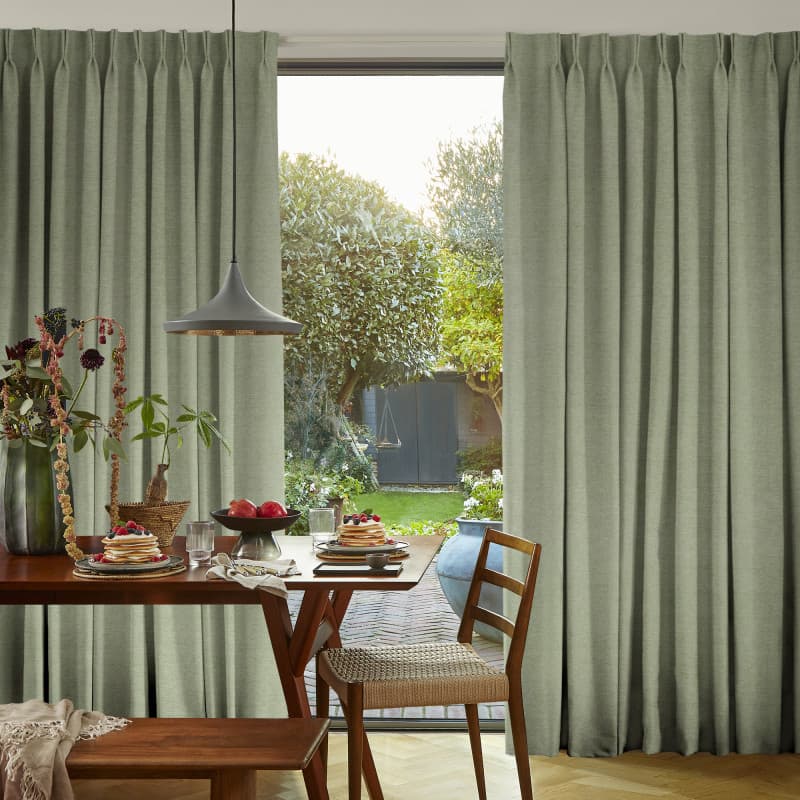 Pleated sage-green drapes, slightly parted at center, framing a sliding glass door; filtering soft daylight onto dining table set with pancakes, pendant lamp, wooden chairs and a leafy garden beyond.