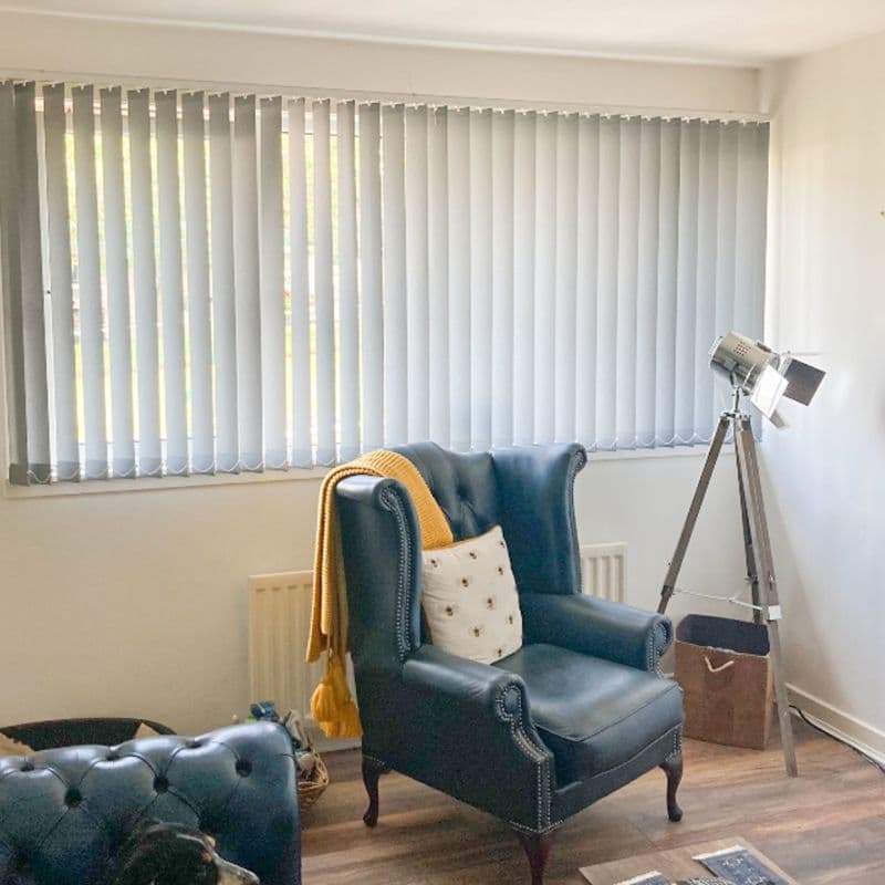 Light-grey vertical blinds cover a wide window, slats drawn and slightly angled, diffusing daylight; bright living room with a dark leather wingback chair draped in a yellow throw and tripod lamp.