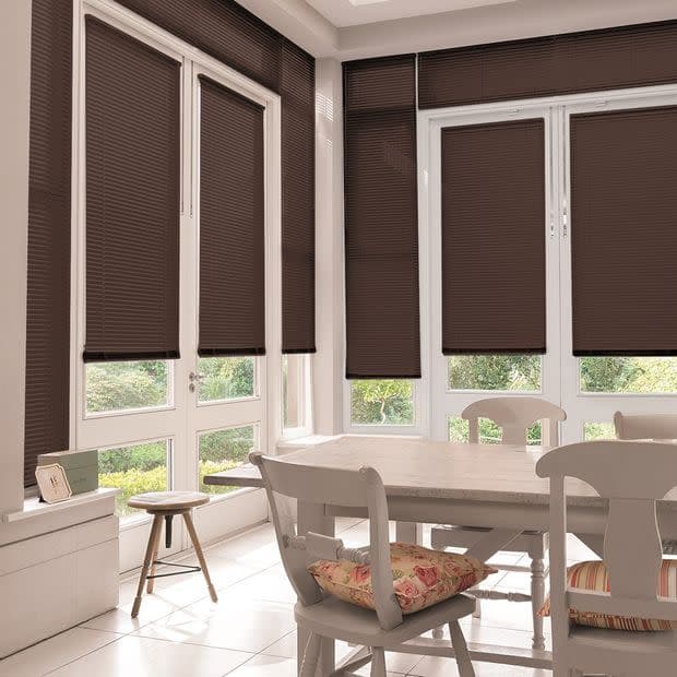 Dark brown pleated blinds covering tall white-framed windows, lowered to block and softly filter daylight; small dining area with white table and chairs, floral cushions and garden view.