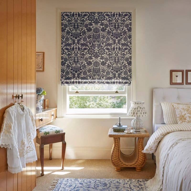 Roman fabric blind with blue floral damask pattern, lowered to cover most of the window and filtering daylight; cozy bedroom setting with bed, wicker bedside table, lamp and wooden wardrobe.