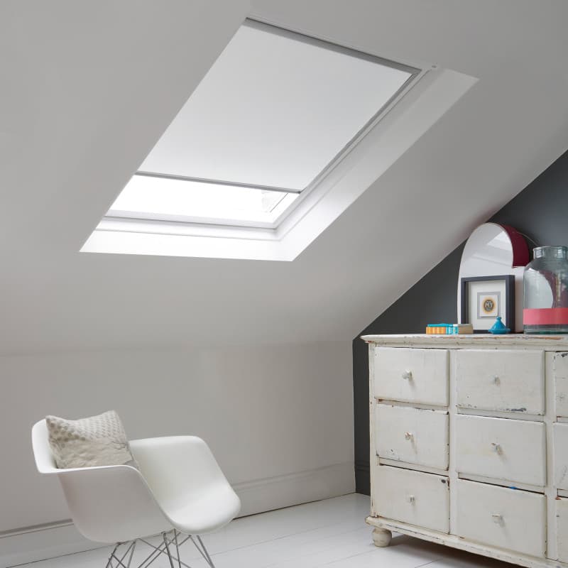 Room with white eaves wall, dark blue-grey wall to right with a shabby chic white drawer unit, a contemporary white chair, white painted floorboards and a skylight blind in white.