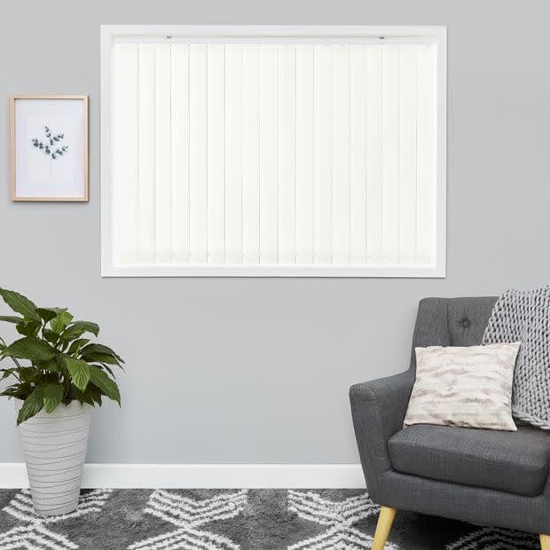 White vertical blinds covering a centered rectangular window, closed and diffusing light; modern living room with grey walls, potted plant left, upholstered grey armchair right and patterned rug.