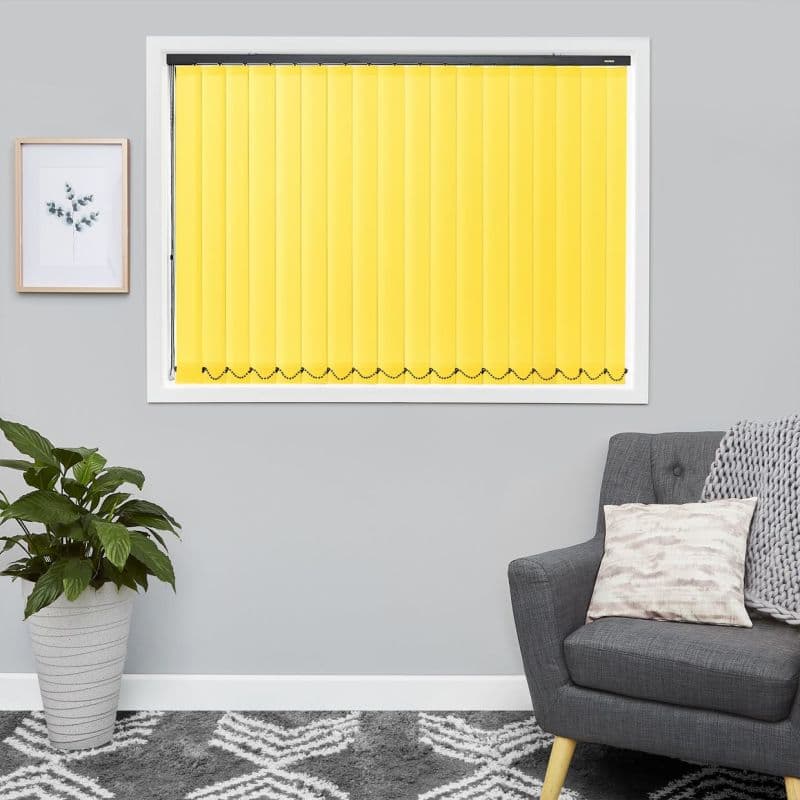 Yellow vertical blinds installed inside a white window frame, fully closed and diffusing light; modern living room with grey walls, framed plant print, potted houseplant, patterned rug and grey armchair.