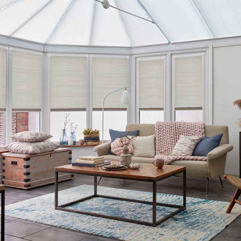 Pleated cellular blinds across tall conservatory windows and glazed roof, beige and fully lowered, diffusing soft daylight; a cozy sitting area with sofa, pink knitted throw, and wooden coffee table.