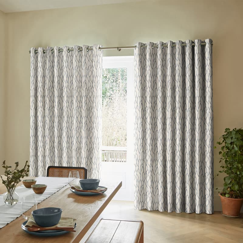 Floor-length grommet-top curtains with pale geometric blue-gray pattern, hung across a sliding glass door, partly drawn to reveal a narrow bright outdoor view; light-filled dining room with table and potted plant.