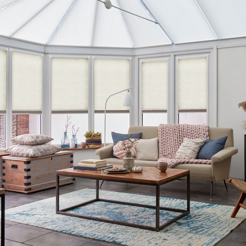 Cellular pleated blinds lowered across tall conservatory windows, gently filtering soft daylight; beige sofa with pink chunky knit throw, wooden coffee table on blue rug, cozy sunroom setting.