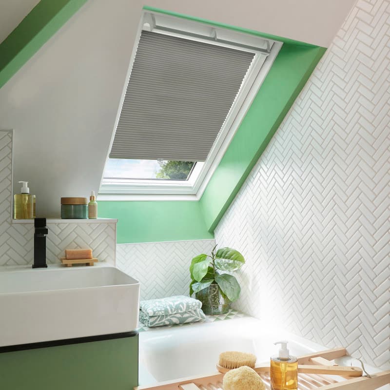 Grey pleated skylight blind, partially lowered and filtering daylight, in a green-accented attic bathroom with white herringbone tiles, sink and bathtub, folded towel and potted plant on ledge.