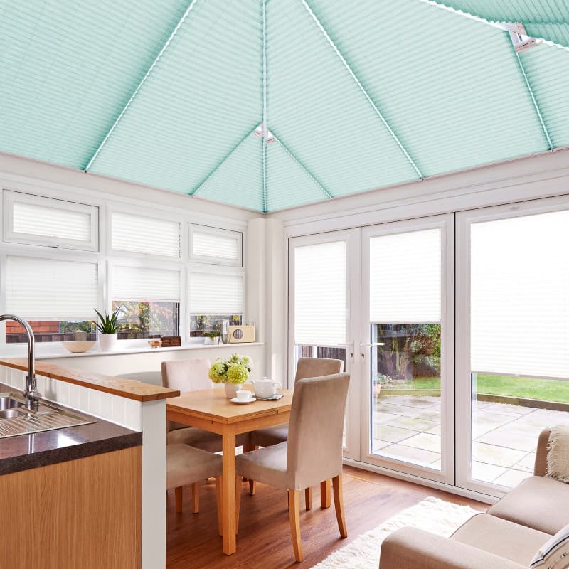 Seafoam pleated roof blinds cover the pitched conservatory ceiling; white pleated blinds cover windows and doors, filtering soft daylight into a bright kitchen-dining conservatory with wooden table and garden view.