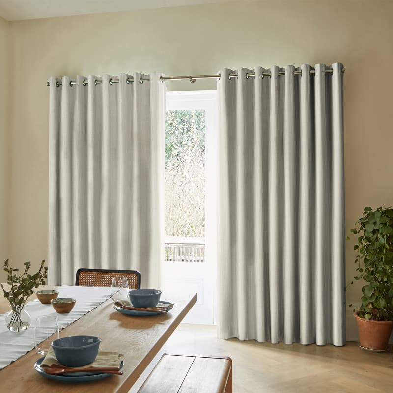 Grommet-top light-gray heavy curtains, floor-length and slightly parted at the center, filtering bright daylight from a glass door; dining room with wooden table settings, bench seating, and a potted plant.