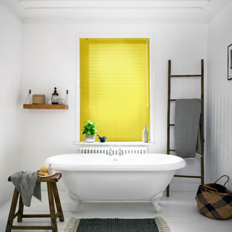 Yellow slatted blinds fully lowered and closed, filtering bright daylight; in a white bathroom with freestanding clawfoot tub, wooden stool, ladder towel rack, woven basket and potted plant.