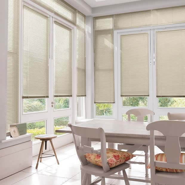 Beige horizontal Venetian blinds covering tall bay windows, lowered and filtering soft daylight; in a bright sunroom dining area with white table, chairs, floral cushions and garden view.