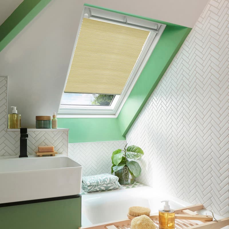 A pleated yellow skylight blind, mounted inside the frame, is partially lowered, filtering daylight over a sloped bathroom with green-painted trim, white herringbone tiles, sink, tub, and plant.