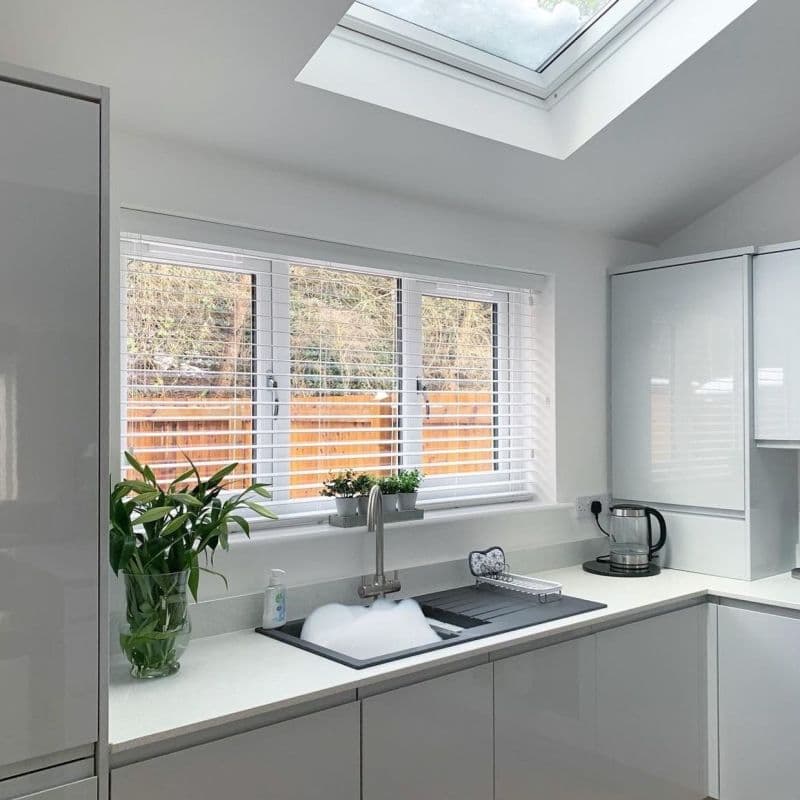 White horizontal Venetian blinds lowered, slats tilted to let light through over three-panel kitchen window above soapy sink, in a bright modern white kitchen with skylight and potted plants.