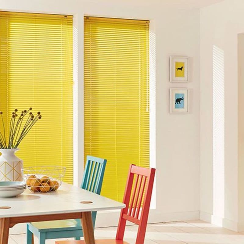 Yellow horizontal Venetian blinds covering two tall adjacent windows, fully lowered with slats closed, filtering sunlight into a bright dining nook with white walls, multicolored chairs, table and vase.