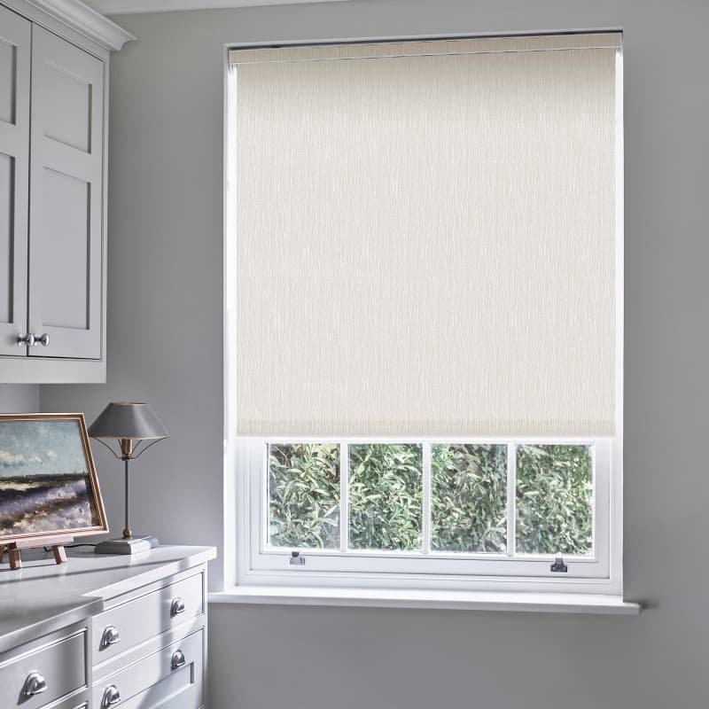 Beige textured roller blind, fully lowered with bottom just above sill, filtering daylight; in a light-gray utility/kitchen with white cabinets, a lamp, painting on the counter, and leafy garden outside.