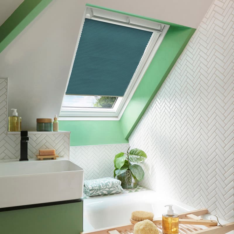 Teal pleated skylight blind, almost fully lowered over a roof window, filtering daylight; attic bathroom setting with green-painted window recess, white herringbone tiles, sink and bathtub with plant and toiletries.
