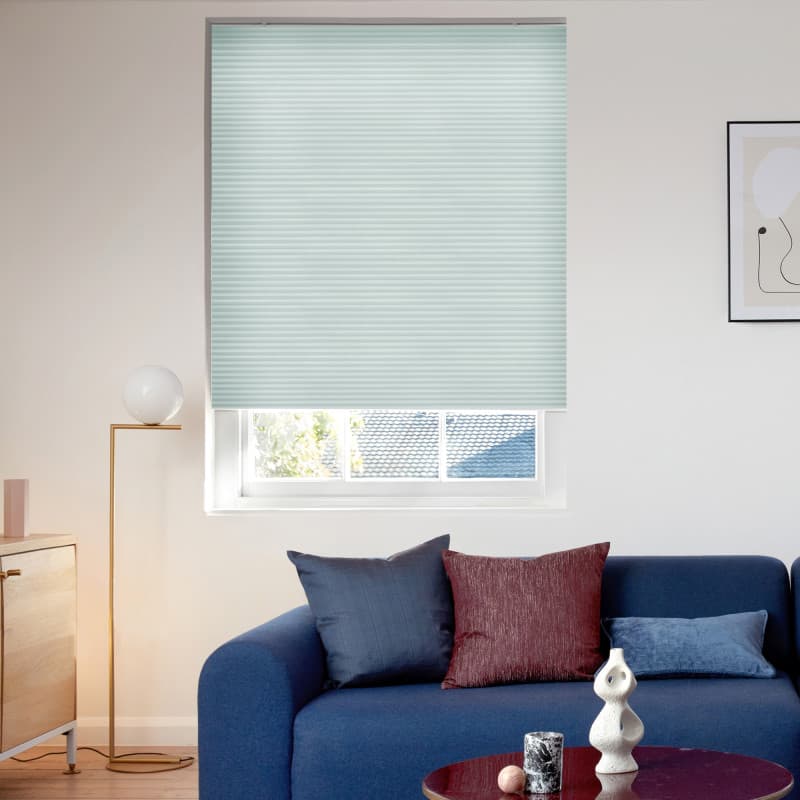 Light-teal cellular blind, lowered with a narrow gap at bottom, filtering soft daylight; modern living room with blue sofa, cushions, floor lamp, coffee table and small roof-and-tree view.