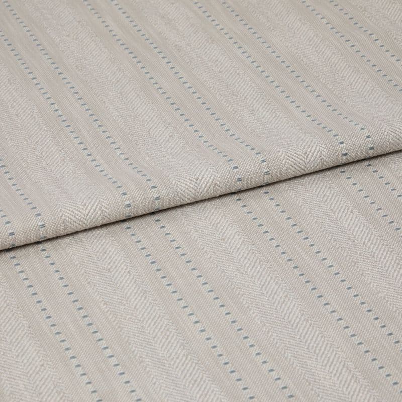 Beige woven fabric featuring herringbone chevrons and thin blue dotted vertical stripes â€” folded over itself, displayed as a close-up textile sample on a neutral, softly lit surface.