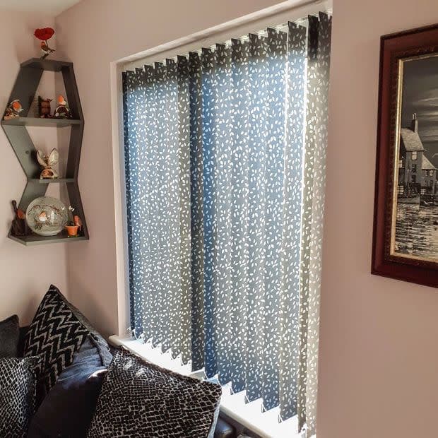 Patterned blue-and-white vertical blinds covering a window, closed and filtering daylight; set in a living-room corner above a sofa with patterned cushions, decorative hexagonal shelf and framed wall art.