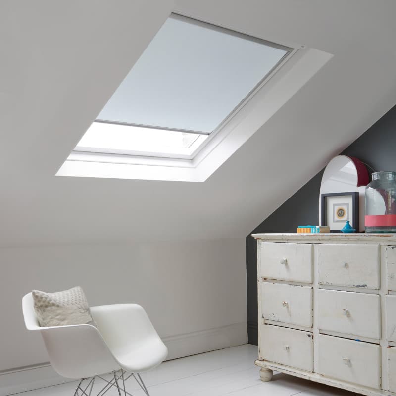 Room with white eaves wall, dark blue-grey wall to right with a shabby chic white drawer unit, a contemporary white chair, white painted floorboards and a skylight blind in pale blue.