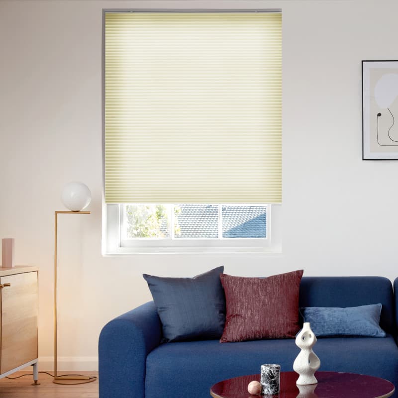 Cellular blind, pale yellow, lowered across a recessed window, filtering light and leaving the lower pane exposed; living room with blue sofa, cushions, floor lamp, wooden sideboard, coffee table.