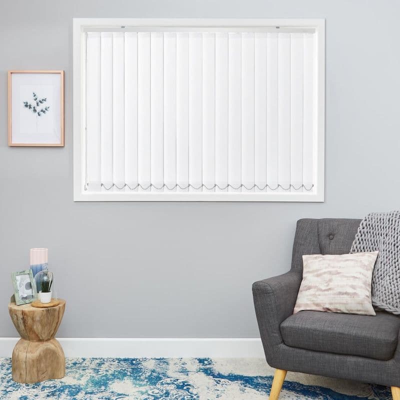 Vertical white blinds mounted inside the window, slatted panels closed and light-filtering with beaded bottom chain; in a gray living room with gray armchair, wooden side table and blue rug.
